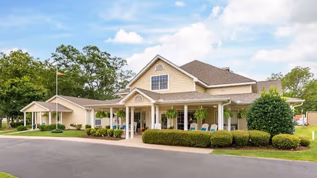 Exterior view of a single-story senior living facility building with beige siding, a gabled roof, a covered porch with white columns, hanging plants, and several chairs. The building is surrounded by neatly trimmed bushes, a small lawn, and trees under a partly cloudy sky.