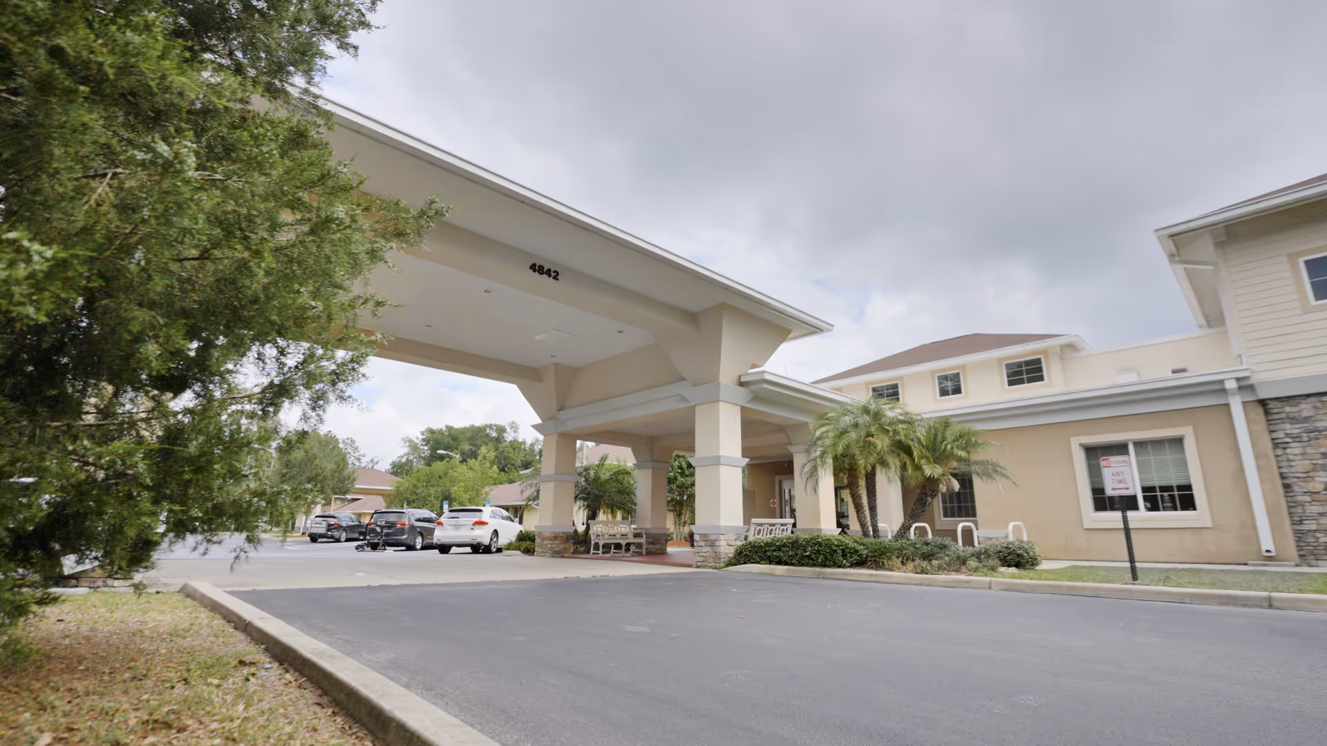 Exterior view of Plaza Health and Rehab Center showing the main entrance with a covered drop-off area, several parked cars, palm trees, and a cloudy sky.