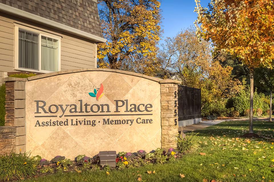 Stone sign for Royalton Place Assisted Living and Memory Care facility, surrounded by green grass, flowers, and trees with autumn foliage, next to a beige building and a black metal fence.
