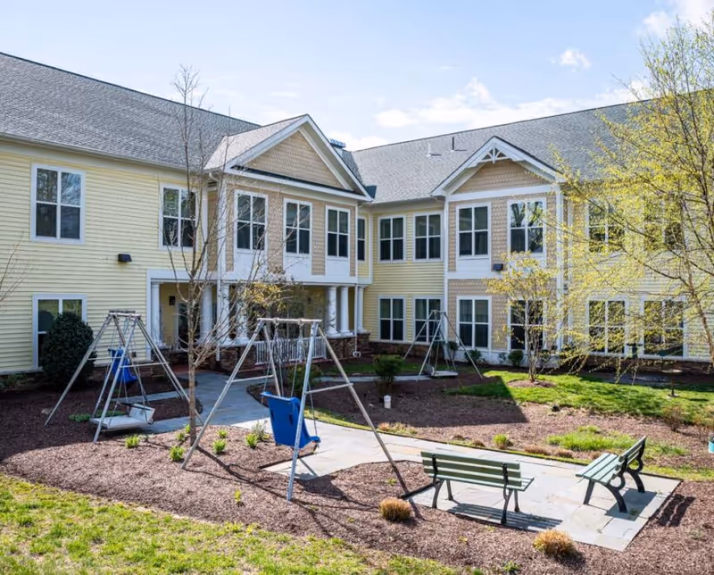 Outdoor courtyard area of a senior living facility with yellow two-story building in the background, featuring multiple windows. The courtyard has a paved walkway, several swings including a blue adaptive swing, benches, young trees, and landscaped garden beds with mulch and grass.
