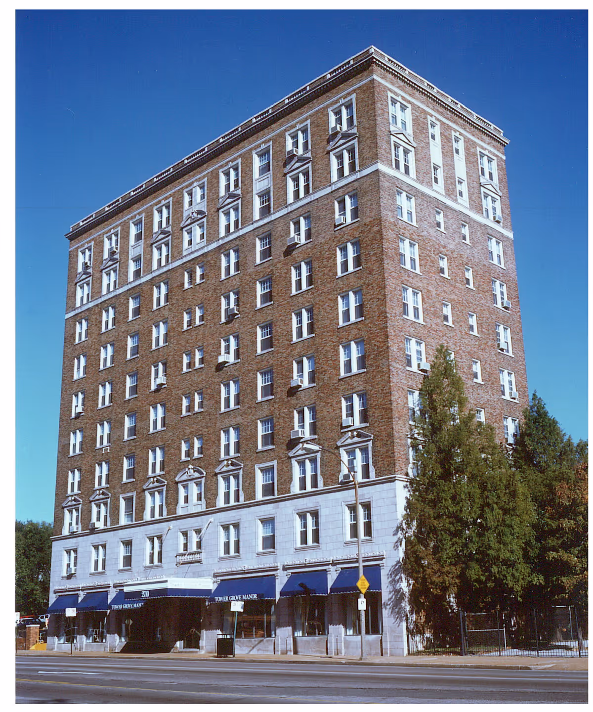 A multi-story brick building with white window frames and decorative architectural details. The building has blue awnings over the entrances at street level, with the name 'Tower Grove Manor' visible on the awnings. There are trees on the right side of the building and a clear blue sky above.