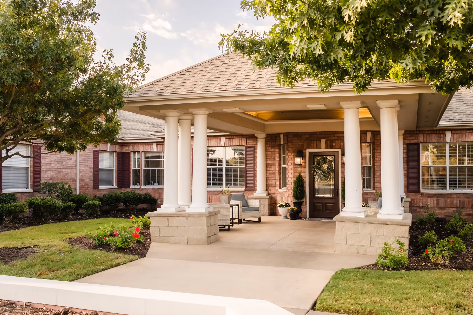 Front entrance of a brick building with a covered porch supported by white columns, surrounded by green grass, bushes, and trees under a partly cloudy sky.