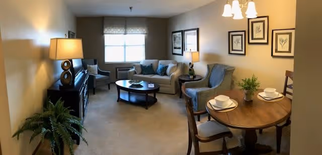A cozy living room and dining area featuring a beige sofa with blue cushions, two armchairs, a round wooden coffee table, a wooden dining table set for two with white plates and bowls, framed botanical artwork on the wall, a floor lamp, and a potted plant near the entrance.