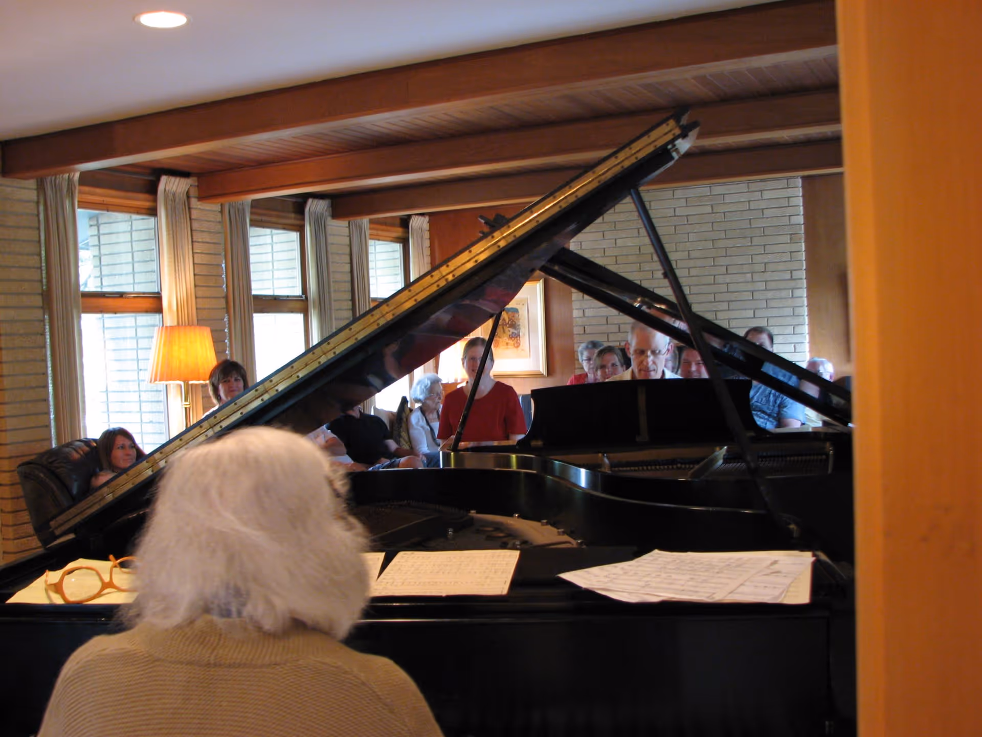 An elderly person with white hair is playing a grand piano in a room with wooden beams and large windows. Several people are seated in the background, attentively watching the performance. Sheet music and glasses are placed on the piano.