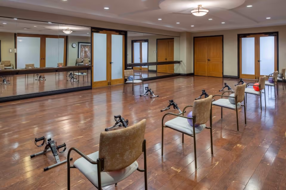 A spacious exercise room with wooden flooring, several chairs arranged in a row each with an exercise ball on the seat, and pedal exercisers placed on the floor in front of the chairs. The room has large mirrors on one wall, wooden double doors, and soft overhead lighting.