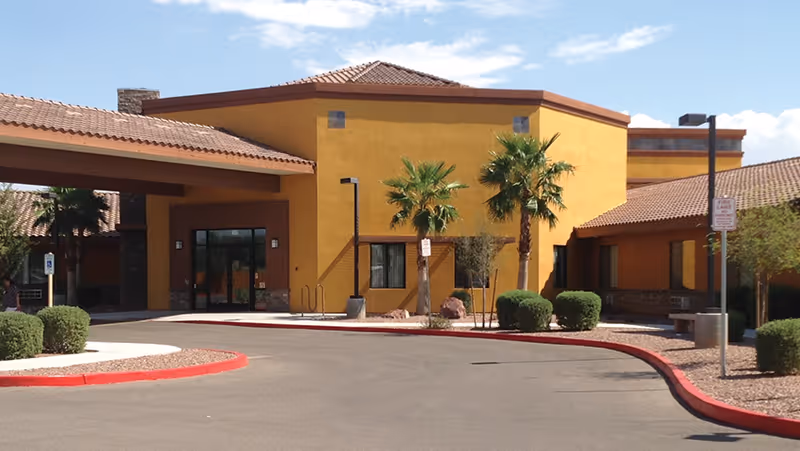 Entrance of a yellow stucco nursing facility with a covered drop-off, palm trees, and a curved red-painted curb.