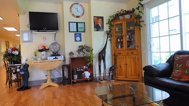 A cozy living room area with wooden flooring, a black leather couch with a colorful pillow, a glass coffee table, a wooden cabinet with plants and decorative items, two giraffe sculptures, a wall-mounted TV, a clock, framed certificates, and a portrait on the wall. There are also various plants and flowers placed around the room, and a large window letting in natural light.