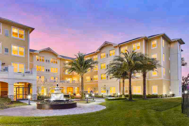Exterior view of Sonata Windermere senior living facility at dusk, showing a multi-story building with many lit windows, palm trees, a central fountain, and a well-maintained lawn and walkway.