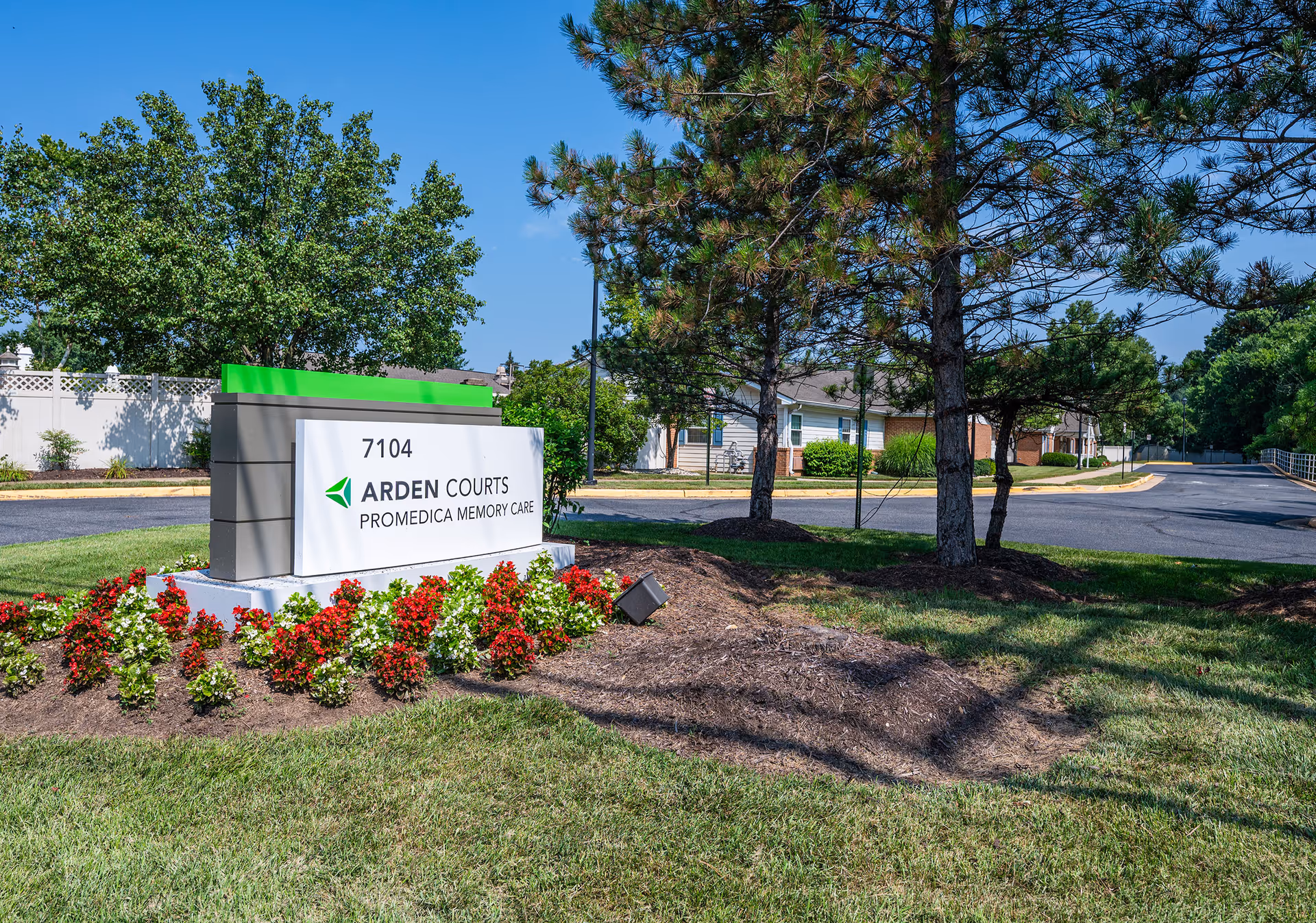 Entrance sign for Arden Courts ProMedica Memory Care with flowers, trees, and the facility driveway in the background.