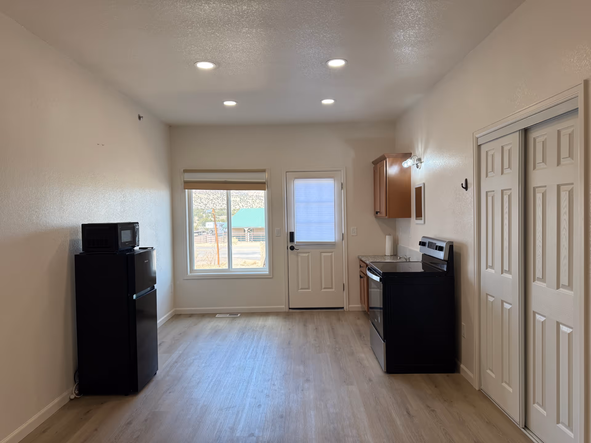 A small kitchen area with a black refrigerator and microwave on the left, a window and a door in the center, and a stove with a cabinet and sink on the right. The room has light-colored walls and wood flooring, with recessed ceiling lights and a closet with sliding doors on the right.