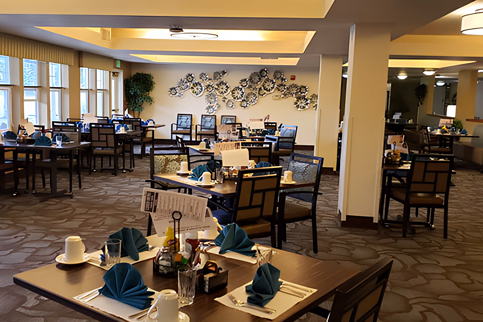 Spacious dining room with multiple set tables, folded napkins, and decorative wall art in a senior living facility.