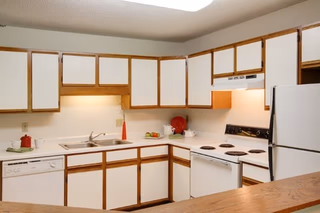 A clean kitchen with white cabinets and wooden trim, featuring a double sink, dishwasher, electric stove with four burners, white refrigerator, and a wooden countertop. There are a few decorative items including a red teapot, a red vase with dried flowers, and a plate with fruit on the counter.