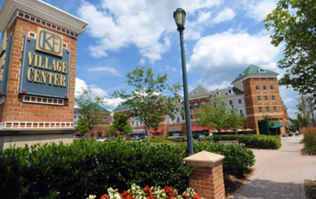 Outdoor view of a village center with a brick sign reading 'Village Center' on the left, surrounded by greenery and flowers. In the background, there are multi-story brick buildings with shops and trees lining a paved walkway under a partly cloudy sky.