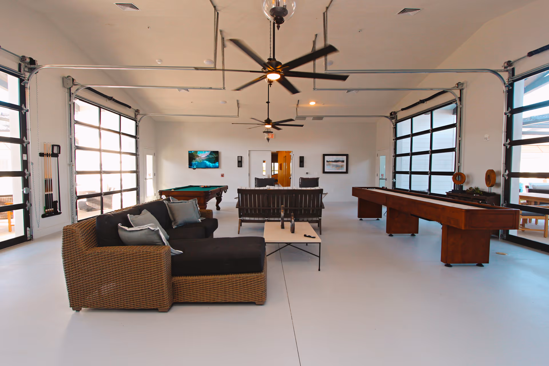 Spacious community game room with a wicker sectional sofa, pool and shuffleboard tables, ceiling fans and large garage-style glass doors.