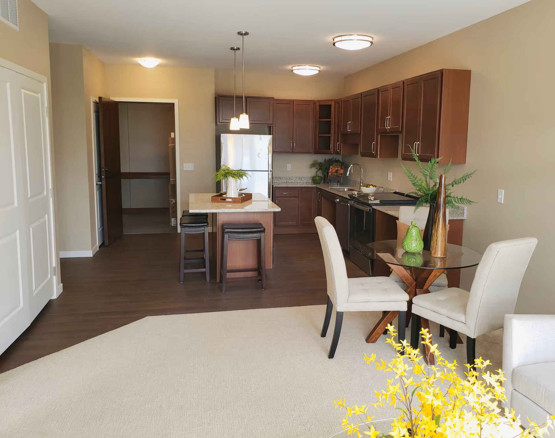 A modern kitchen and dining area in a senior living facility. The kitchen features dark wood cabinets, a stainless steel refrigerator, stove, and dishwasher. There is a small island with two stools and pendant lighting above it. Adjacent to the kitchen is a round glass dining table with two upholstered chairs. The room has beige walls and a mix of wood and carpet flooring. Decorative plants and a yellow flower arrangement add color to the space.