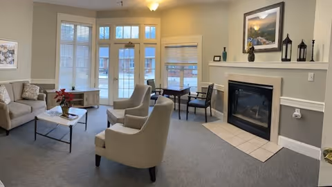 A cozy senior living facility common area with a fireplace, two beige armchairs, a beige sofa, a coffee table with a poinsettia plant, a side table, and a small desk with two chairs near large windows with blinds.