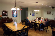 A group of elderly people sitting around a table in a well-lit dining room with wooden floors and light green walls. The room has multiple wooden tables and chairs, pendant lights hanging from the ceiling, and framed artwork on the walls.