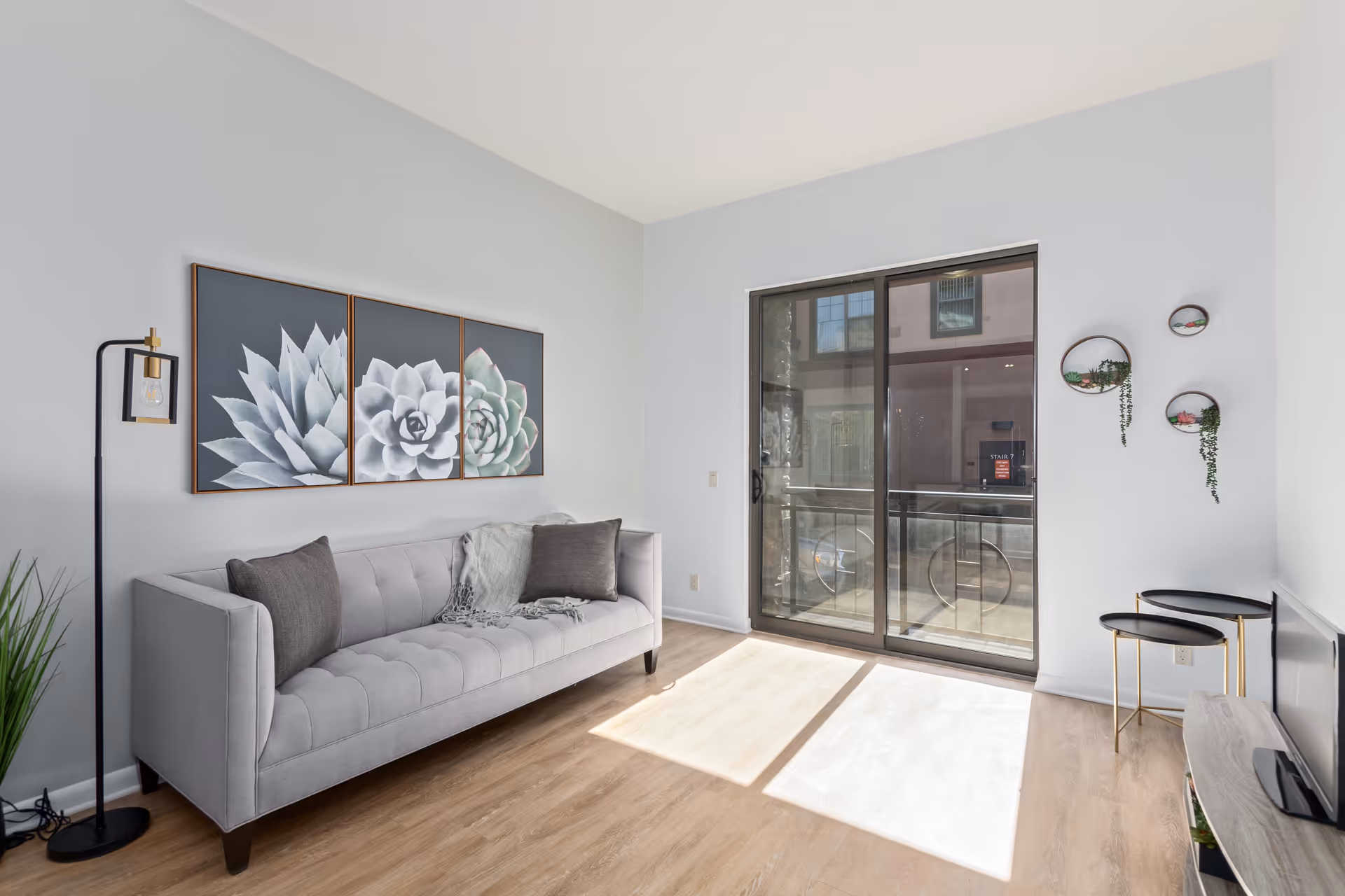 A bright living room with a light gray tufted sofa adorned with two dark gray pillows and a gray throw blanket. Above the sofa are three framed succulent plant artworks. To the left of the sofa is a modern floor lamp with a visible bulb. On the right side of the room, there are three small round wall-mounted planters with green and pink plants. A sliding glass door lets in natural light and leads to an outdoor balcony. The floor is light wood, and a small TV is visible on a stand in the corner.