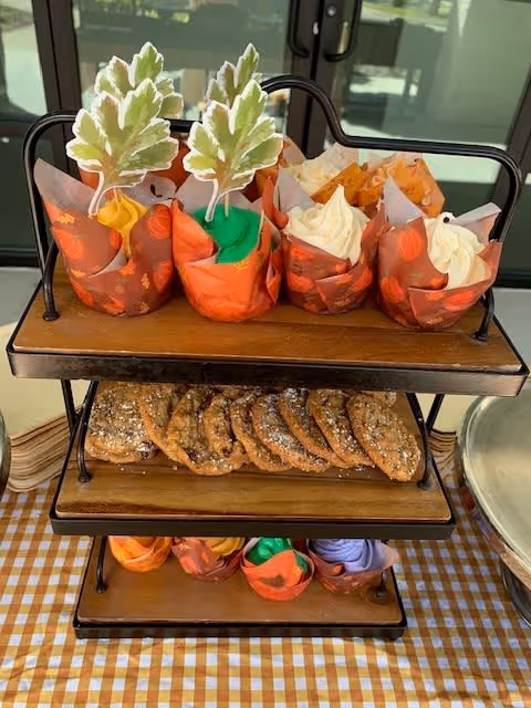 A three-tiered wooden and metal stand displaying fall-themed cupcakes decorated with leaf-shaped toppers and cookies on a checkered tablecloth in front of glass doors.