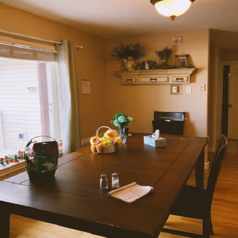 Dining room with a large wooden table and chairs, baskets and a tissue box on the table, and a window with curtains.