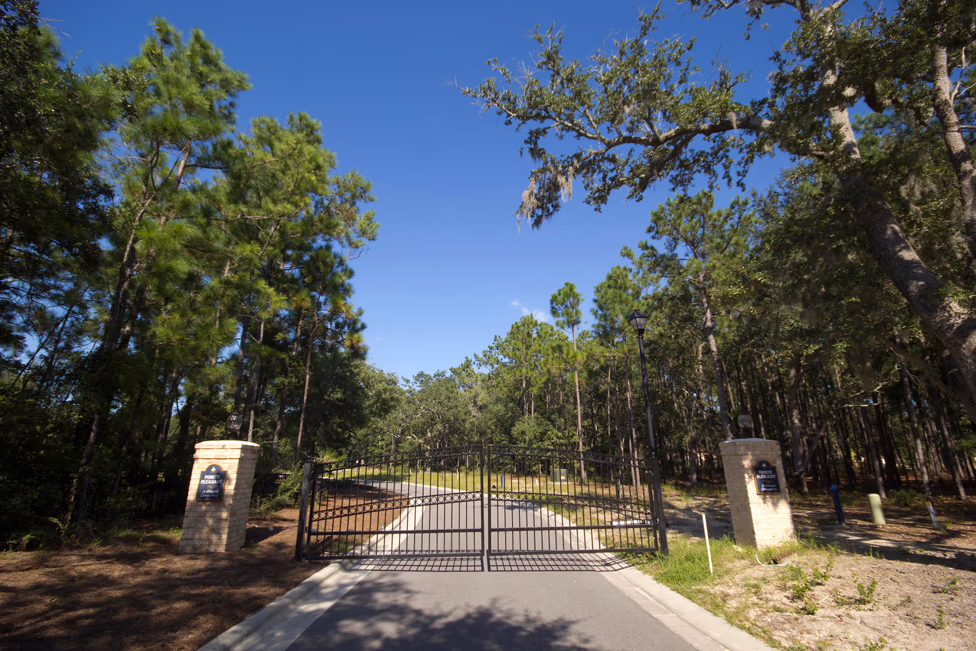A gated entrance to a facility surrounded by tall pine trees and greenery under a clear blue sky. The entrance features a black metal gate flanked by two stone pillars with signs on them.