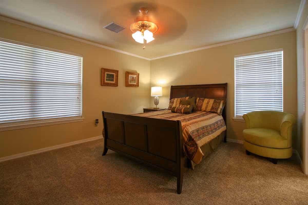A cozy bedroom with a wooden bed frame, a patterned bedspread, and several pillows. The room has beige walls, two windows with white blinds, a ceiling fan with lights, a small nightstand with a lamp, two framed pictures on the wall, and a green upholstered armchair in the corner.