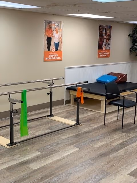 A physical therapy or rehabilitation room with parallel bars for walking exercises, a padded therapy table with cushions, and a chair. The room has wood-patterned flooring, beige walls with white wainscoting, and two motivational posters on the wall. One poster reads 'Greater Independence' and the other reads 'Once Thought Impossible.'