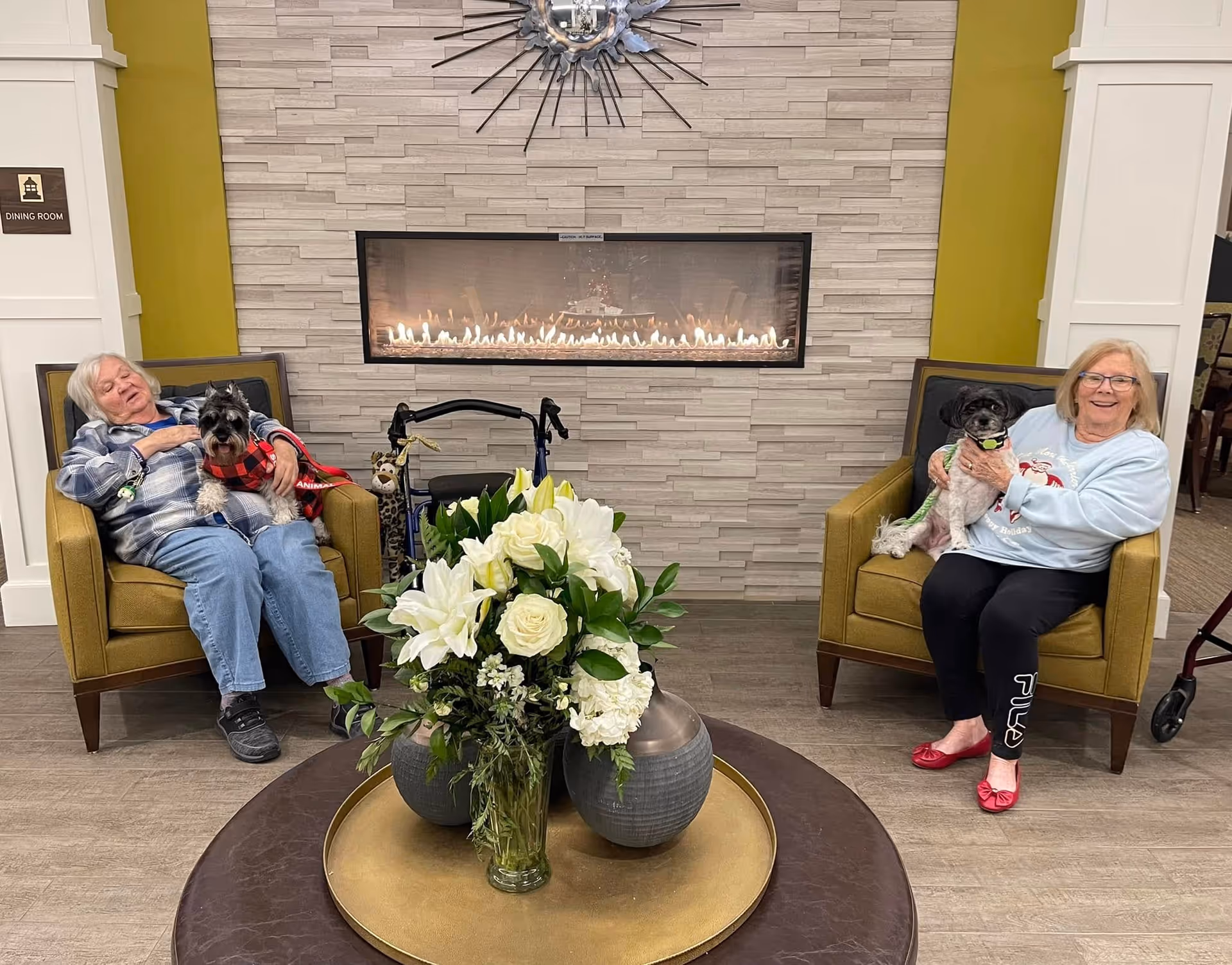 Two elderly women sitting in armchairs in a cozy room with a modern fireplace on the wall behind them. Each woman is holding a small dog. There is a round table with a large floral arrangement in the foreground. A walker is positioned between the chairs, and a sign on the wall indicates the dining room nearby.
