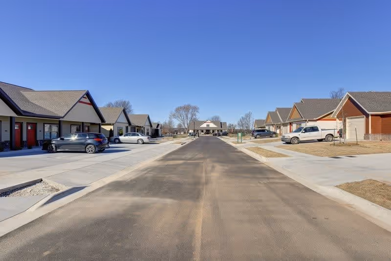 A paved road flanked by single-story residential buildings with attached garages and parked cars on both sides under a clear blue sky.