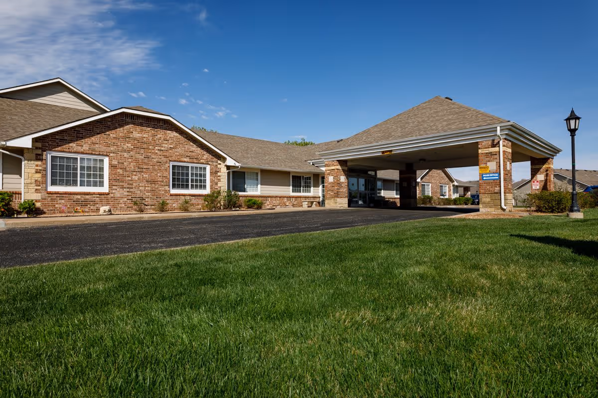 Exterior view of Homestead Estates of Wichita showing a single-story brick building with a covered entrance driveway, green lawn in the foreground, and a clear blue sky.
