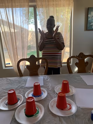 A person standing behind a table with six red plastic cups placed upside down on white paper plates, each plate having a colored ring underneath the cup. The person is silhouetted against a window with sheer curtains, and there are wooden chairs around the table.