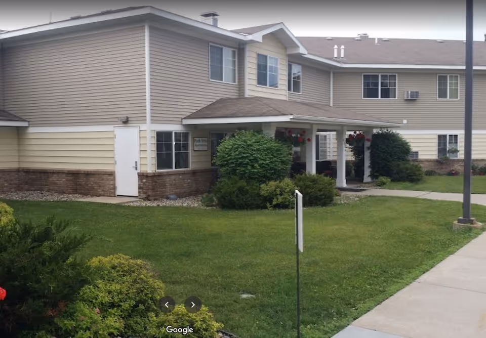 Exterior front entrance of a two-story apartment building with a covered porch, lawn, and shrubs.