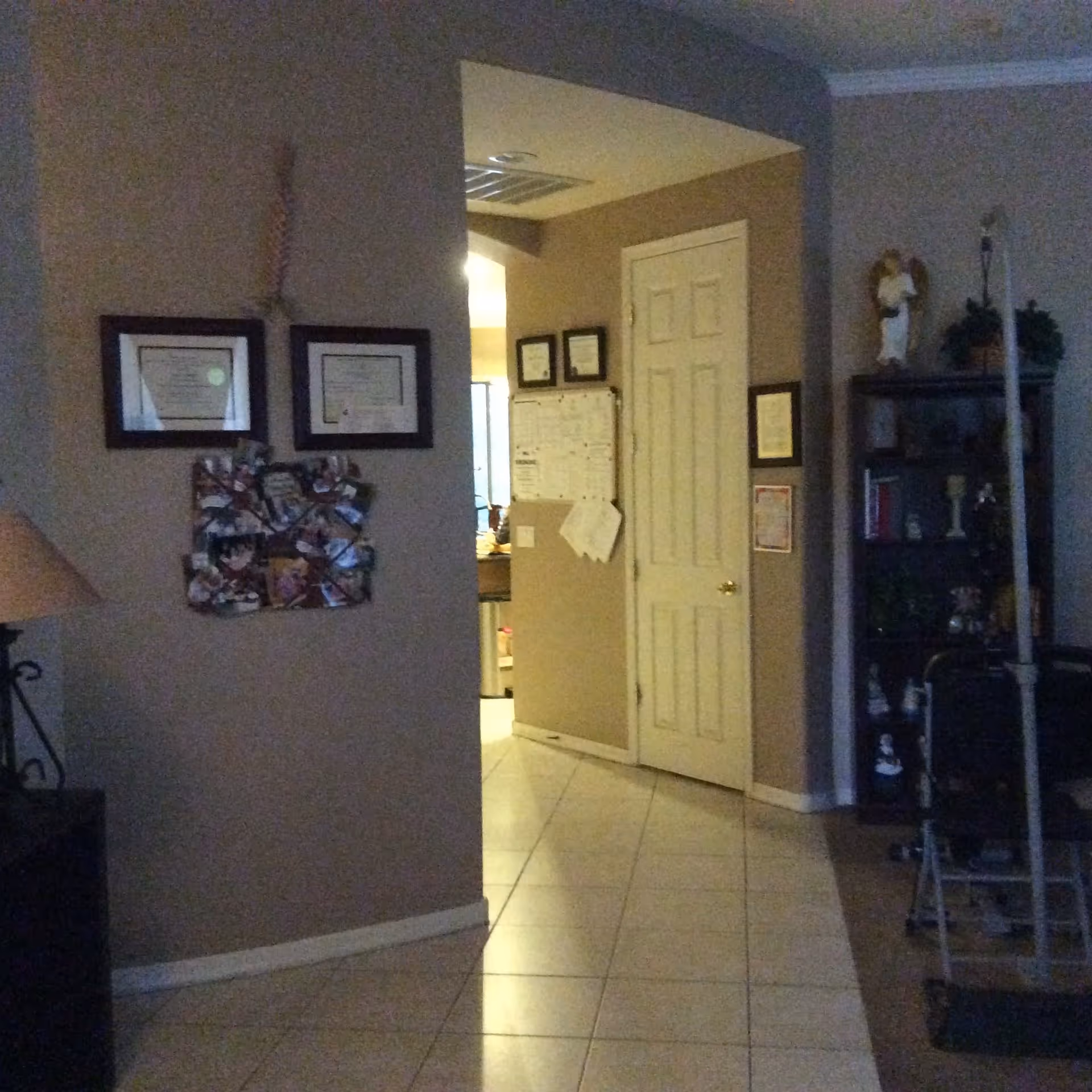 Interior view of a living space with beige walls and tiled floor. There are framed certificates and a photo collage on the left wall, a white door in the center, and a dark wooden shelf with decorative items and plants on the right. A lamp is partially visible on the left side, and some exercise equipment is on the right side of the image.