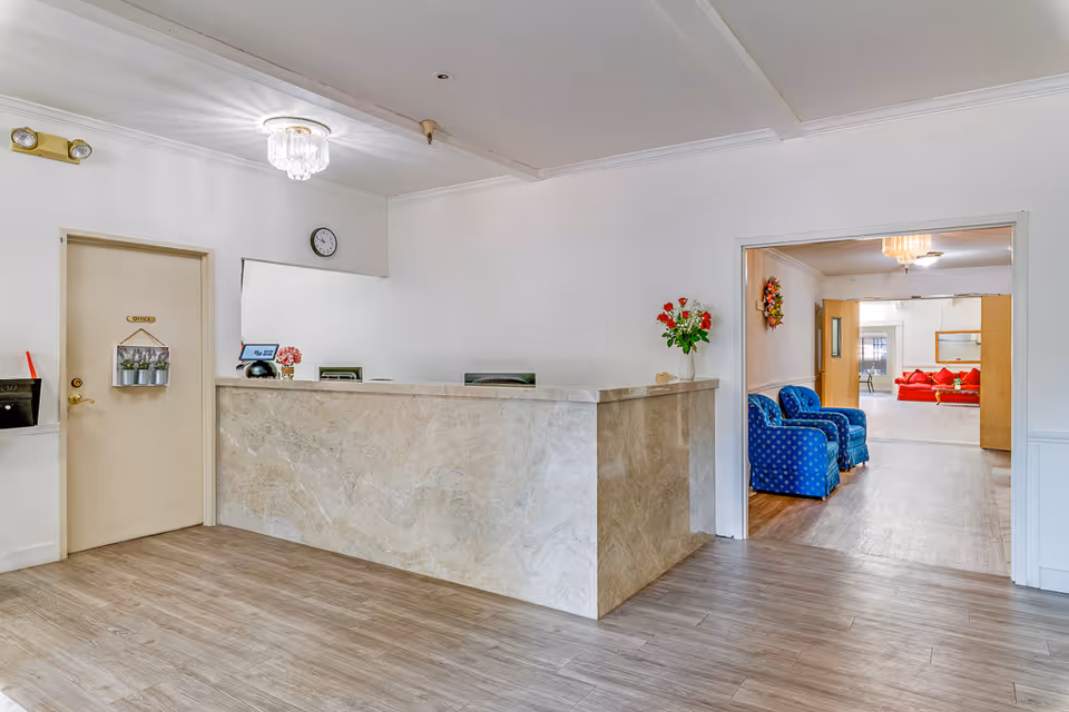 Reception area of Leisure Vale Assisted Living with a marble front desk, a door labeled 'Office' on the left, a clock on the wall, and a vase with red and white flowers on the desk. Through an open doorway, a seating area with two blue armchairs and a red sofa is visible.