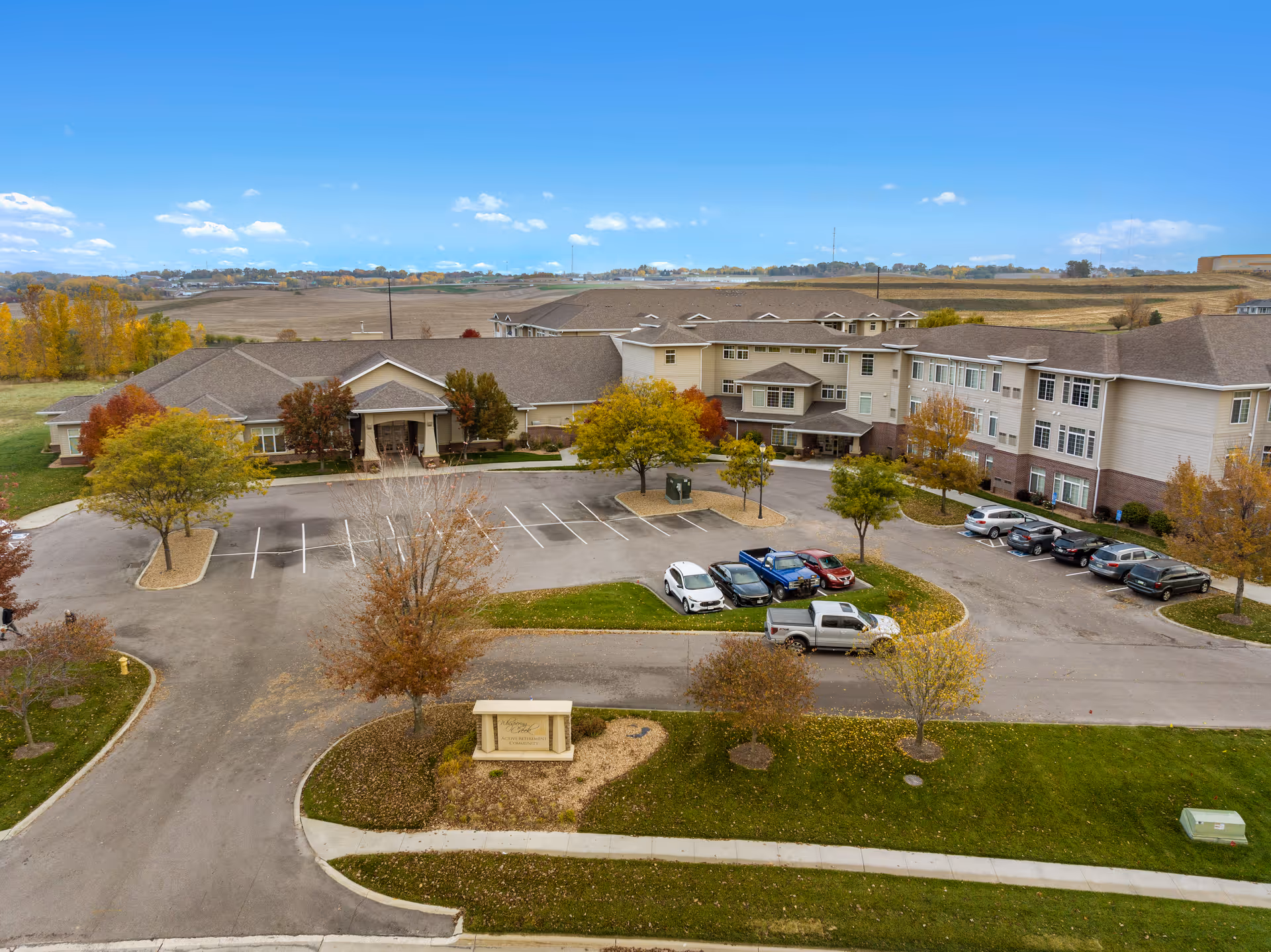 Aerial view of Whispering Creek Senior Living facility showing a large multi-story building with a parking lot in front. The building is surrounded by trees with autumn foliage and open fields in the background under a clear blue sky.