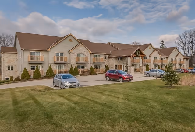 Front view of a two-story senior living facility with balconies, a parking lot, and a landscaped lawn under a partly cloudy sky.