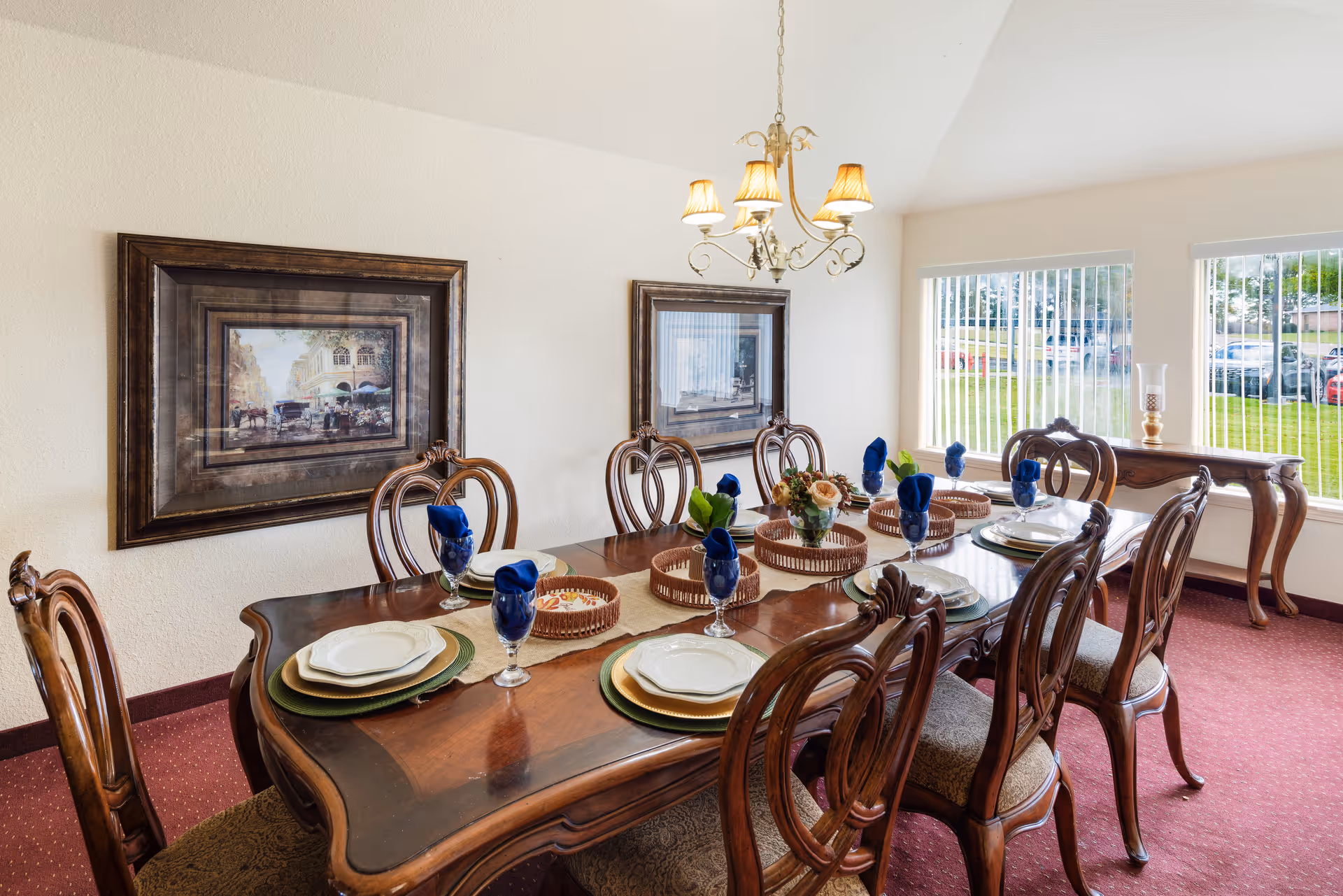 A formal dining room with a polished wooden table set for eight people. The table has white plates, green placemats, blue napkins in glasses, and decorative baskets and a floral centerpiece. The room has two framed paintings on the wall, a chandelier with five lampshades hanging above the table, and large windows with vertical blinds letting in natural light. A wooden side table with a candle holder is positioned against the wall near the windows.