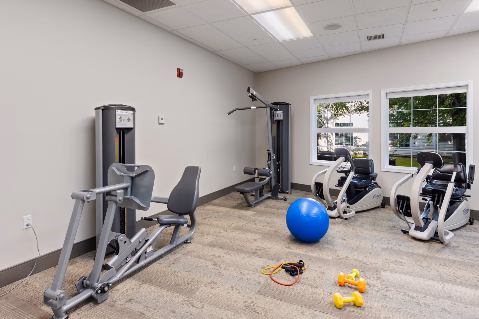A senior living facility exercise room with various fitness equipment including two recumbent exercise bikes, a multi-function weight machine, a blue exercise ball, resistance bands, and small dumbbells. The room has beige walls, carpeted flooring, and two windows showing greenery outside.