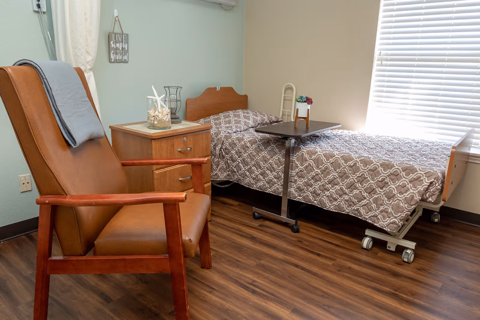 A small, cozy bedroom in a healthcare facility featuring a single hospital-style bed with patterned bedding, a brown leather armchair with a gray blanket draped over the back, a wooden nightstand with decorative items including a glass jar filled with seashells, and a window with white blinds letting in natural light.