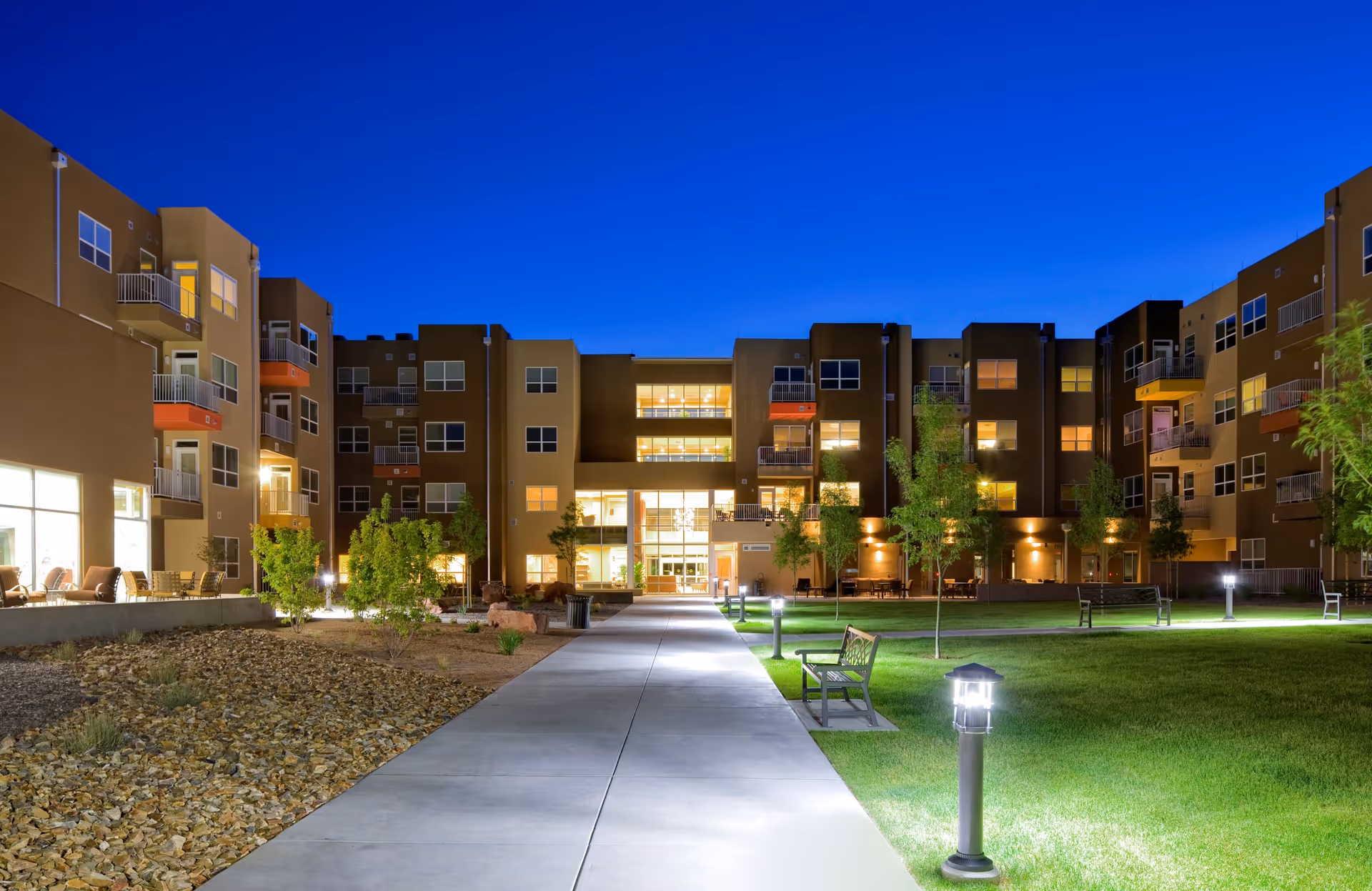 Night view of a modern multi-story senior living facility with well-lit windows and balconies. A wide concrete pathway leads through a landscaped courtyard with green grass, trees, benches, and outdoor lighting fixtures.