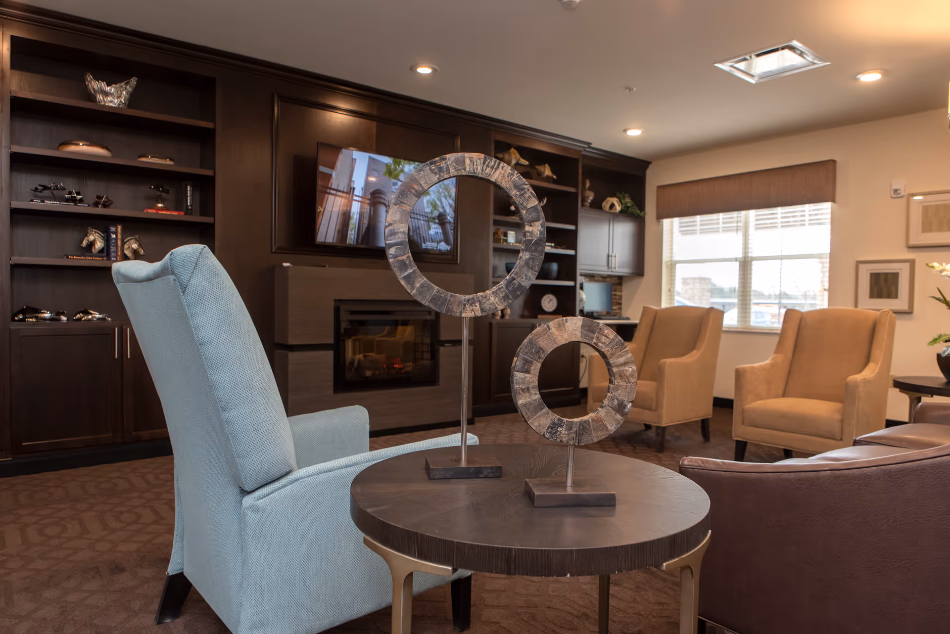 A cozy living room area with a light blue armchair, two beige armchairs, and a brown leather chair arranged around a round wooden table with two decorative circular sculptures. In the background, there is a dark wood built-in shelving unit with a fireplace and a mounted TV above it. A window with blinds lets in natural light.