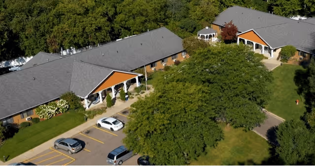 Aerial view of Vibrant Life Senior Living Kalamazoo showing two single-story buildings with gray roofs and orange gable accents, surrounded by green trees and lawns. There is a parking lot with several cars in front of the buildings and a white gazebo visible between the buildings.