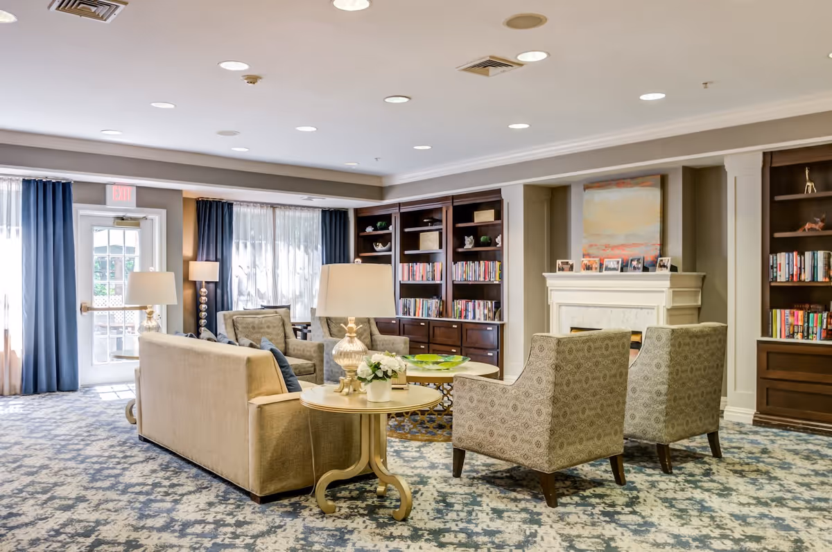 A bright and spacious living room area with a beige sofa, two patterned armchairs, and a round coffee table in the center. There are two side tables with lamps and a vase with flowers. The room features built-in dark wood bookshelves filled with books and decorative items, a white fireplace with framed photos on the mantel, and large windows with sheer and dark blue curtains allowing natural light to enter.