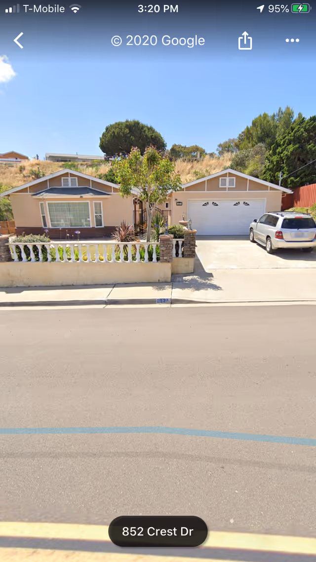 Front exterior view of a single-story house with a beige facade, a large window, a white garage door, a driveway with a parked silver SUV, a small front yard with plants and a tree, and a low white fence with stone pillars.