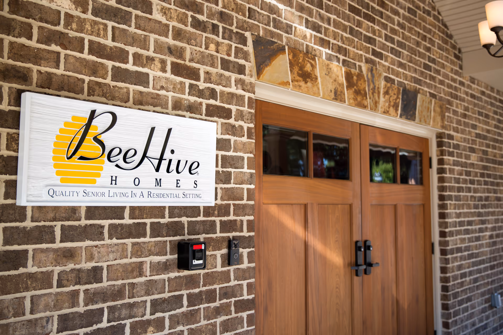 Entrance of BeeHive Homes senior living facility showing a brick wall with a white sign featuring the BeeHive Homes logo and slogan, next to double wooden doors with small windows at the top.