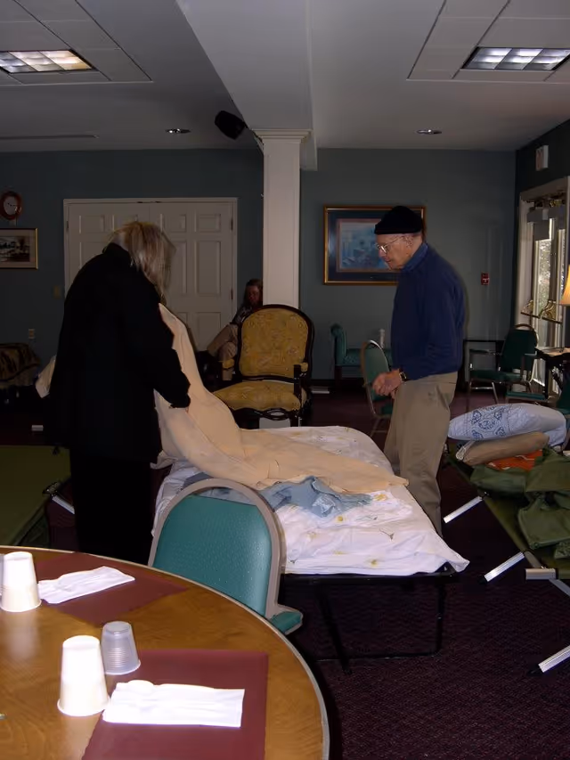 Two elderly individuals in a common room setting, one folding a beige blanket on a cot bed while the other stands nearby. The room has several chairs, a round table with disposable cups and napkins, and framed artwork on the walls.