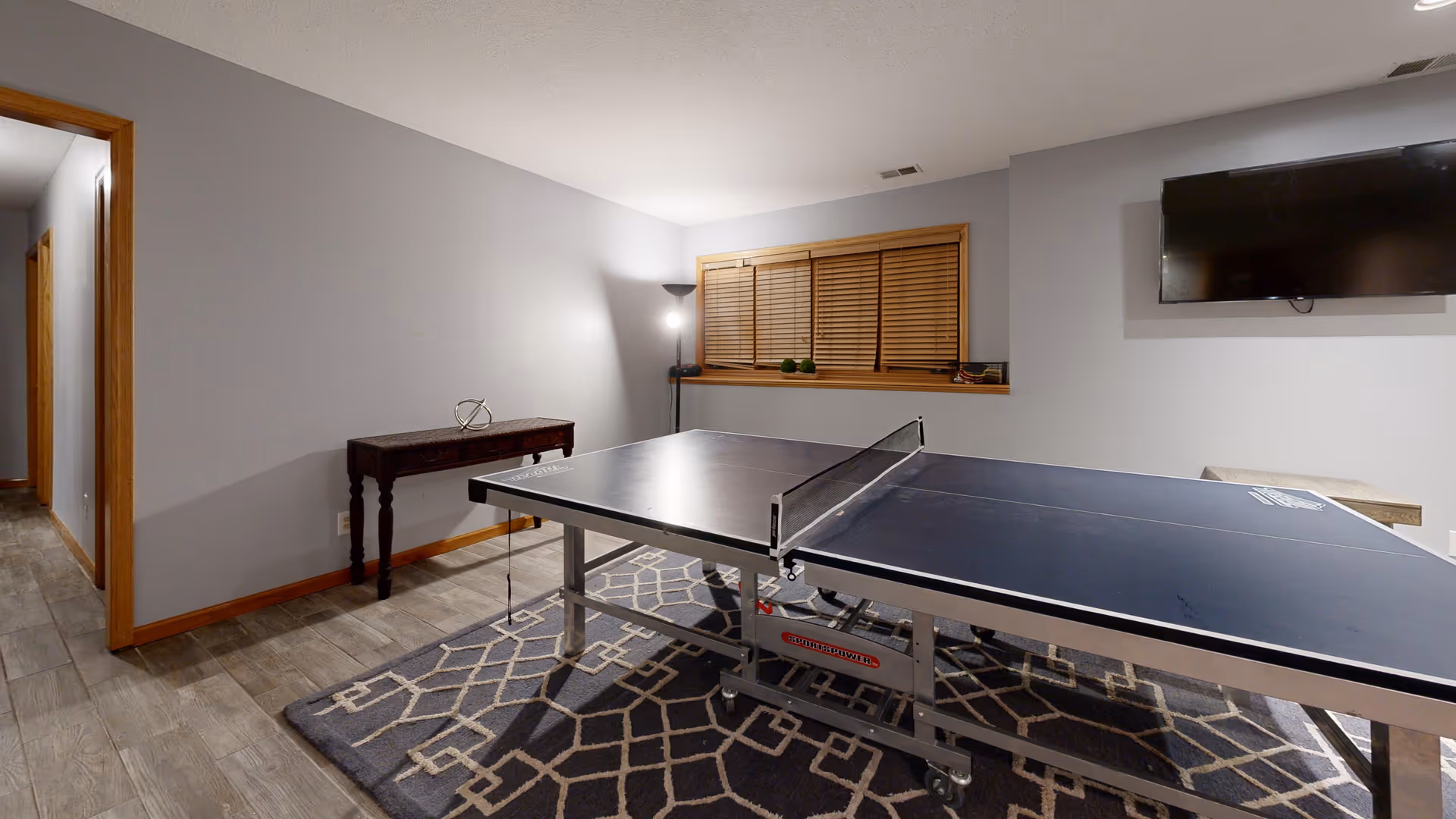 A recreational room with a ping pong table placed on a patterned rug. The room has light gray walls, a window with wooden blinds, a wall-mounted TV, a small table with decorative items, and a floor lamp in the corner. The floor is tiled with a wood-like finish.
