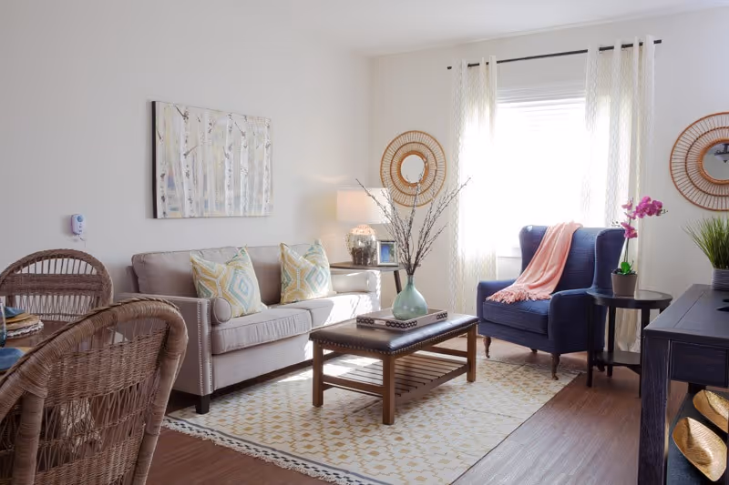 A bright and cozy living room with a beige sofa adorned with patterned cushions, a dark blue armchair with a pink throw blanket, a wooden coffee table with a decorative vase, and a side table with a lamp and picture frame. The room has light-colored walls, a large window with sheer curtains, two round wicker wall mirrors, and a patterned area rug on wooden flooring.