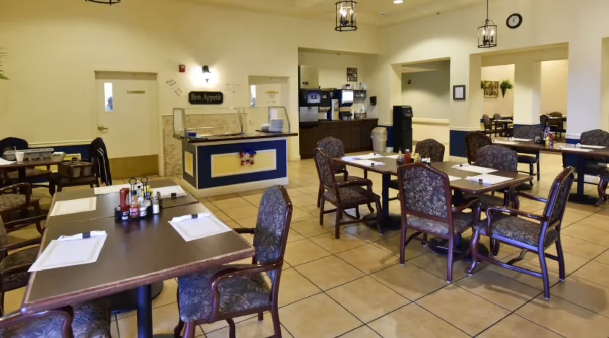 Dining room with several set tables and upholstered chairs, a central serving counter labeled "Bon Appetit" and a beverage station in back.