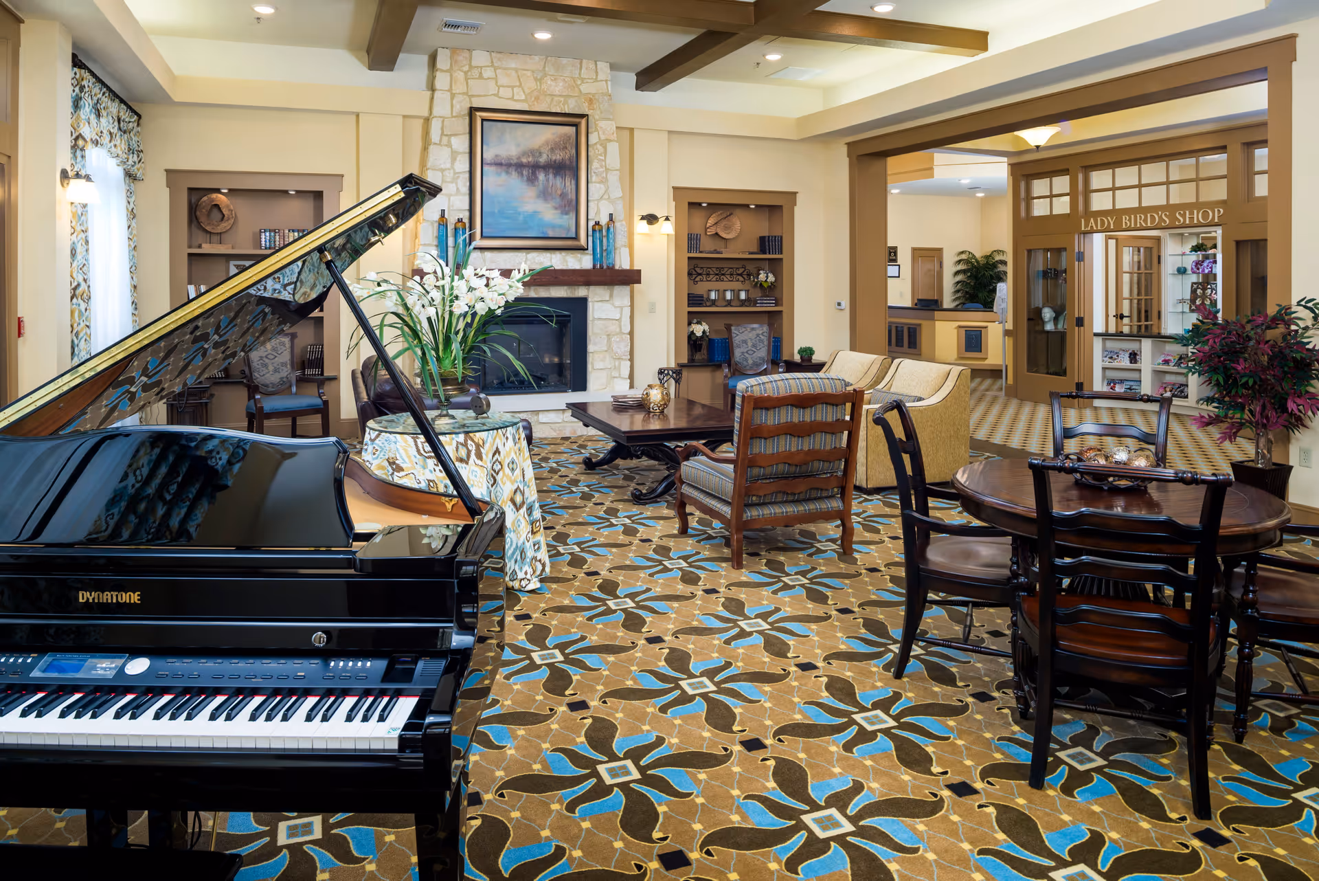 A spacious and well-decorated living room area in a senior living facility featuring a black grand piano, a stone fireplace with a framed painting above it, comfortable seating including armchairs and a sofa, a round wooden table with chairs, decorative plants, and a patterned carpet. In the background, there is an entrance to a shop labeled 'Lady Bird's Shop'.